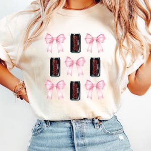 May include: A white t-shirt with a pattern of pink bows and black Coca-Cola Zero cans.