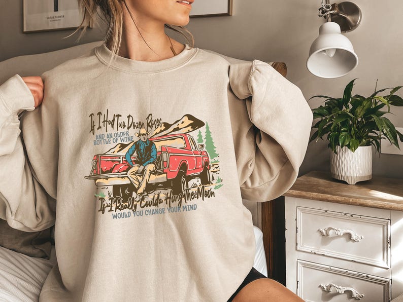 May include: Beige sweatshirt with a vintage-style graphic of a red pickup truck and a cowboy. The text on the sweatshirt reads "If I Had Two Dozen Roses and an Older Bottle of Wine If I Really Coulda Hung The Moon Would You Change Your Mind".