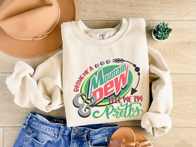 Puede incluir: Una sudadera de color crema con un dise&ntilde;o gr&aacute;fico que presenta el logotipo de Mountain Dew y el texto "Bring me a Mountain Dew & tell me I'm pretty".