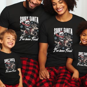 May include: A family of four wearing matching black t-shirts with a graphic of a raccoon riding a reindeer and the text "Sorry Santa I've been Feral". The adults are wearing red and black checked pyjama trousers.