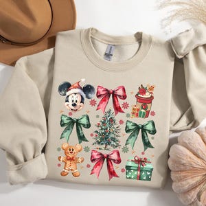 May include: A beige sweatshirt with a Christmas-themed design featuring Mickey Mouse, a gingerbread man, a Christmas tree, bows, and a cup of coffee. The design is in watercolor style with red, green, and brown colors.