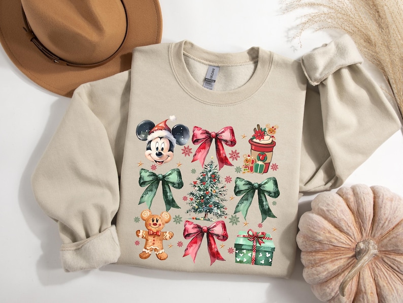 May include: A beige sweatshirt with a Christmas-themed design featuring Mickey Mouse, a gingerbread man, a Christmas tree, bows, and a cup of coffee. The design is in watercolor style with red, green, and brown colors.