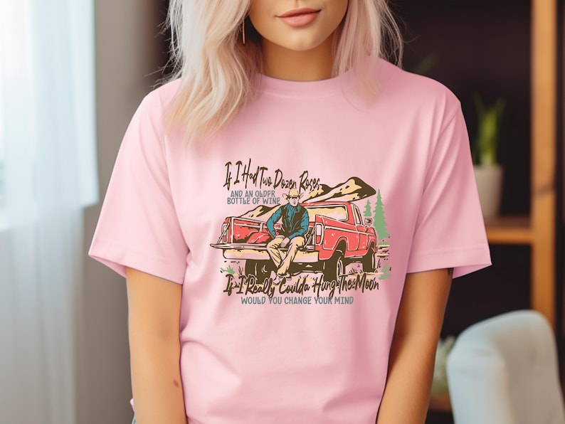May include: Pink t-shirt with a graphic of a man sitting in the back of a red pickup truck. The text on the shirt reads "If I Had Two Dozen Roses and an Older Bottle of Wine If I Really Coulda Hung The Moon Would You Change Your Mind".