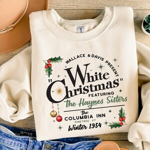 May include: A cream-colored sweatshirt with a vintage-style graphic design featuring the text "Wallace & Davis Present "White Christmas" Featuring The Haynes Sisters The Columbia Inn Pinetree, VT Winter 1954". The graphic also includes holly, stars, and ornaments.