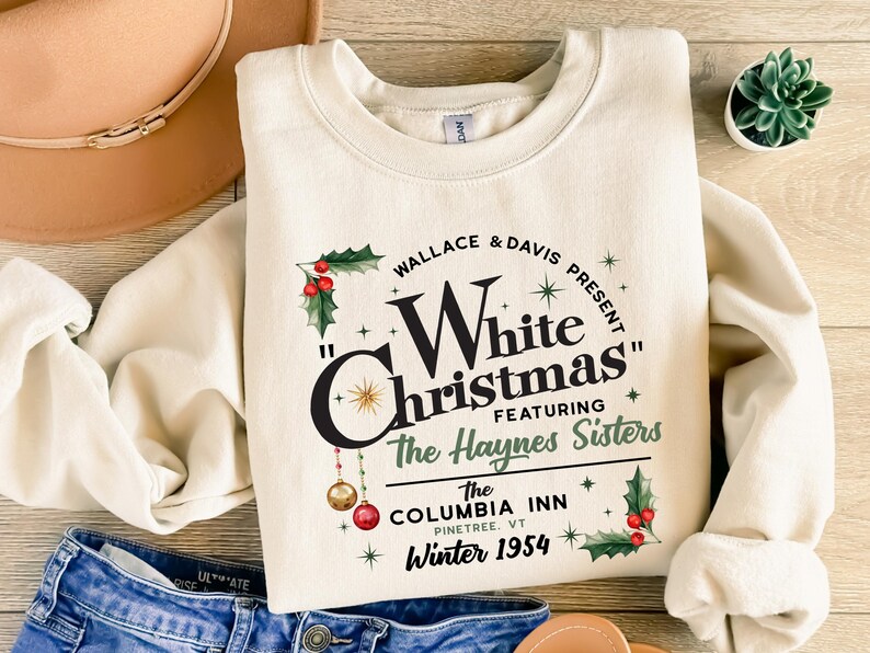 May include: A cream-colored sweatshirt with a vintage-style graphic design featuring the text "Wallace & Davis Present "White Christmas" Featuring The Haynes Sisters The Columbia Inn Pinetree, VT Winter 1954". The graphic also includes holly, stars, and ornaments.