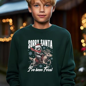 May include: A green Christmas jumper featuring a white graphic of a raccoon wearing a Santa hat riding a reindeer. The text on the jumper reads "Sorry Santa I've been Feral."