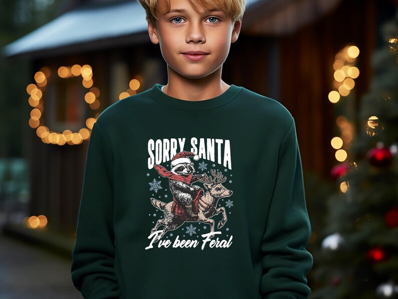 May include: A green Christmas jumper featuring a white graphic of a raccoon wearing a Santa hat riding a reindeer. The text on the jumper reads "Sorry Santa I've been Feral."