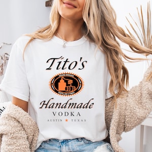 Puede incluir: Camiseta blanca con un gráfico de Tito's Handmade Vodka. El gráfico incluye el logotipo de la marca, que representa un alambique estilizado y barriles, y el texto "Award Winning, Distilled 5 Times, Tito's Handmade Vodka, Austin, Texas".
