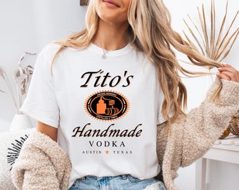 Op de afbeelding: Wit T-shirt met een afbeelding van Tito's Handmade Vodka. De afbeelding bevat het logo van het merk, dat een gestileerde distilleerketel en vaten toont, en de tekst "Award Winning, Distilled 5 Times, Tito's Handmade Vodka, Austin, Texas".