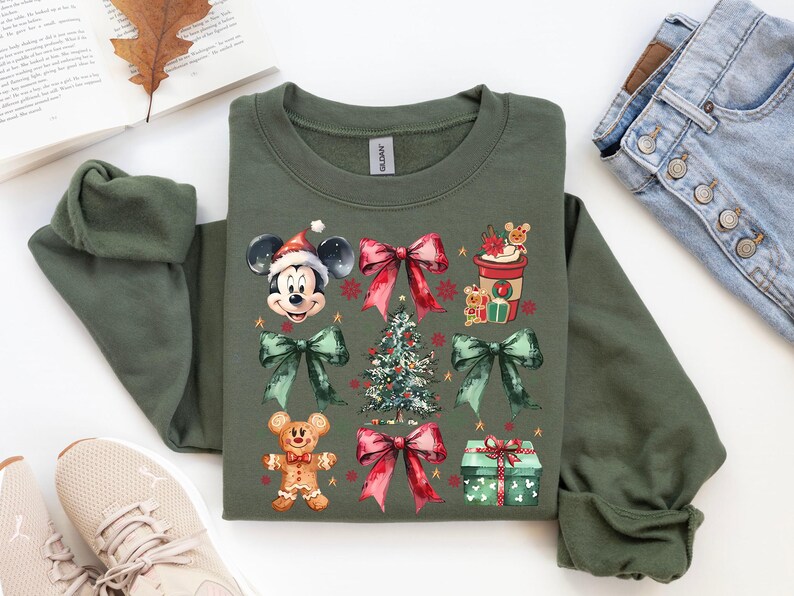 May include: A green sweatshirt with a Christmas-themed design featuring Mickey Mouse, a gingerbread man, a Christmas tree, and a cup of hot cocoa. The design is in a watercolor style with red, green, and gold accents.