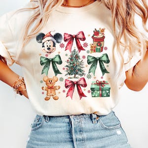 May include: A white t-shirt with a Christmas-themed design featuring Mickey Mouse, a gingerbread man, a Christmas tree, presents, and bows. The design is in a watercolor style with red, green, and brown colors.