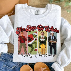 Puede incluir: Una sudadera gris con un gráfico navideño con los personajes de las películas 'Solo en casa', 'El Grinch' y 'Elf'. El gráfico dice 'The Boys of Winter' y 'Merry Christmas'.