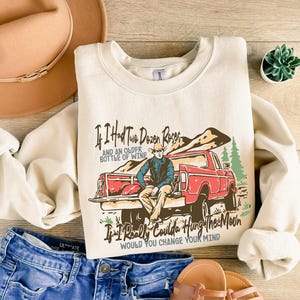 May include: A cream-colored sweatshirt with a vintage-style graphic of a red pickup truck and a man in a cowboy hat. The text on the sweatshirt reads "If I had two dozen roses and an older bottle of wine, if I really coulda hung the moon, would you change your mind?"