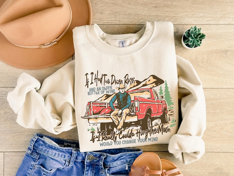 May include: A cream-colored sweatshirt with a vintage-style graphic of a red pickup truck and a man in a cowboy hat. The text on the sweatshirt reads "If I had two dozen roses and an older bottle of wine, if I really coulda hung the moon, would you change your mind?"