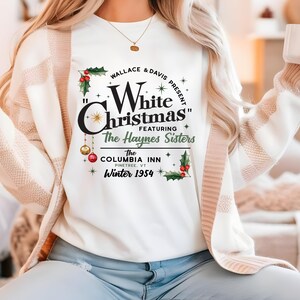 May include: White long-sleeved shirt with a graphic print of a vintage Christmas movie poster. The poster features the text "Wallace & Davis Present "White Christmas" Featuring The Haynes Sisters The Columbia Inn Pinetree, VT Winter 1954".