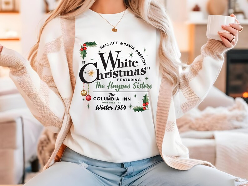 May include: White long-sleeved shirt with a graphic print of a vintage Christmas movie poster. The poster features the text "Wallace & Davis Present "White Christmas" Featuring The Haynes Sisters The Columbia Inn Pinetree, VT Winter 1954".