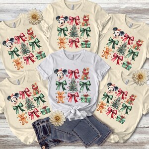 May include: Five cream-colored t-shirts with a Christmas theme featuring Mickey Mouse, a Christmas tree, presents, and bows. The shirts are arranged in a circle with a pair of blue jeans in the center.