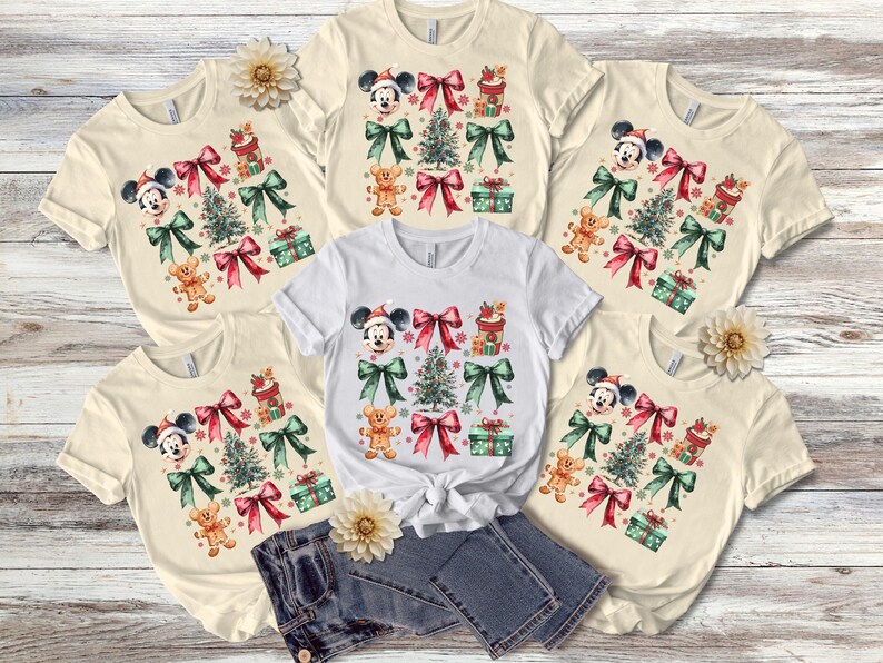 May include: Five cream-colored t-shirts with a Christmas theme featuring Mickey Mouse, a Christmas tree, presents, and bows. The shirts are arranged in a circle with a pair of blue jeans in the center.
