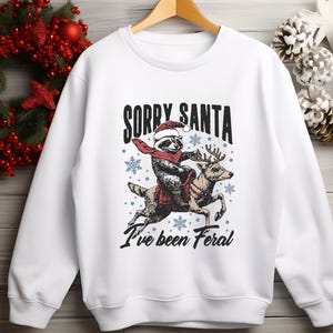 May include: White Christmas sweatshirt featuring a graphic of a raccoon wearing a Santa hat riding a reindeer. The text on the sweatshirt reads "Sorry Santa I've been Feral."