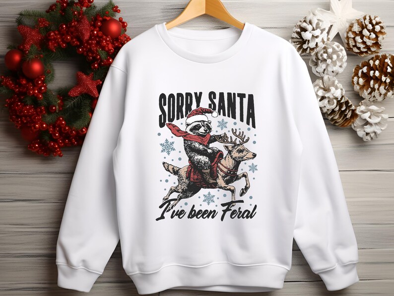May include: White Christmas sweatshirt featuring a graphic of a raccoon wearing a Santa hat riding a reindeer. The text on the sweatshirt reads "Sorry Santa I've been Feral."