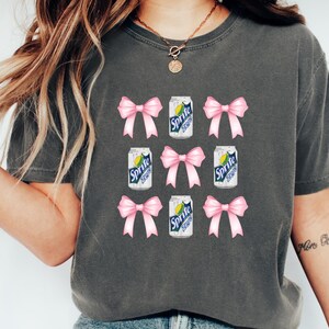 Op de afbeelding: Een grijs T-shirt met een herhalend patroon van roze strikken en blikjes Sprite. De blikjes hebben de tekst "Sprite Zero" erop.