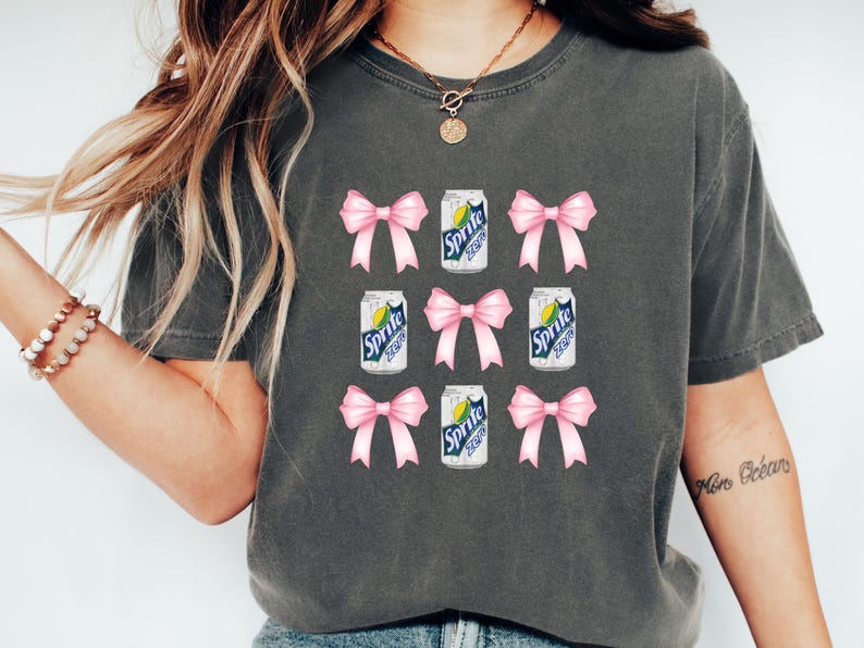 Op de afbeelding: Een grijs T-shirt met een herhalend patroon van roze strikken en blikjes Sprite. De blikjes hebben de tekst "Sprite Zero" erop.