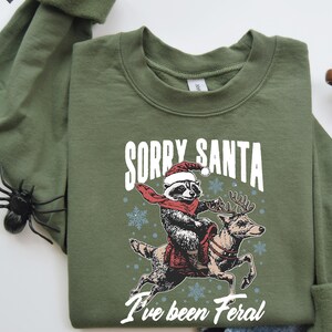 May include: A green sweatshirt featuring a graphic of a raccoon wearing a Santa hat riding a reindeer. The text "Sorry Santa I've been Feral" is printed on the sweatshirt.