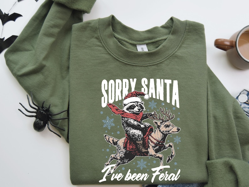 May include: A green sweatshirt featuring a graphic of a raccoon wearing a Santa hat riding a reindeer. The text "Sorry Santa I've been Feral" is printed on the sweatshirt.