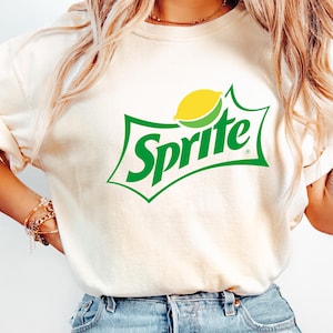 Puede incluir: Una camiseta blanca con un logotipo de Sprite verde y amarillo. El logotipo presenta la palabra "Sprite" en letras verdes con una rodaja de limón amarilla.