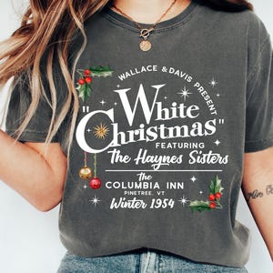 May include: A gray t-shirt with a white graphic design that says "Wallace & Davis Present "White Christmas" Featuring The Haynes Sisters The Columbia Inn Pinetree, VT Winter 1954". The graphic design includes holly, stars, and ornaments.