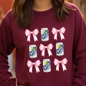 Op de afbeelding: Een maroon sweatshirt met een herhalend patroon van roze strikken en blikjes Sprite Zero. De blikjes zijn wit met groene en gele accenten en de tekst "Sprite Zero" in zwart.