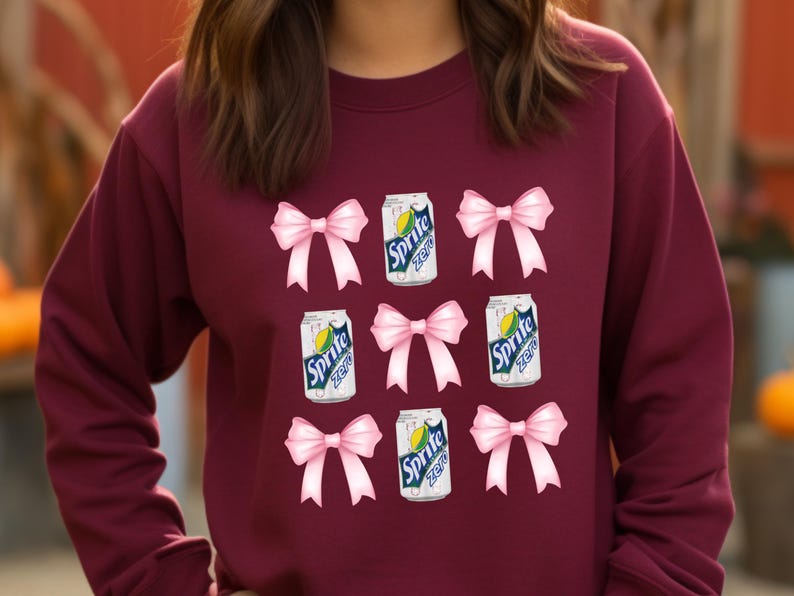 Op de afbeelding: Een maroon sweatshirt met een herhalend patroon van roze strikken en blikjes Sprite Zero. De blikjes zijn wit met groene en gele accenten en de tekst "Sprite Zero" in zwart.