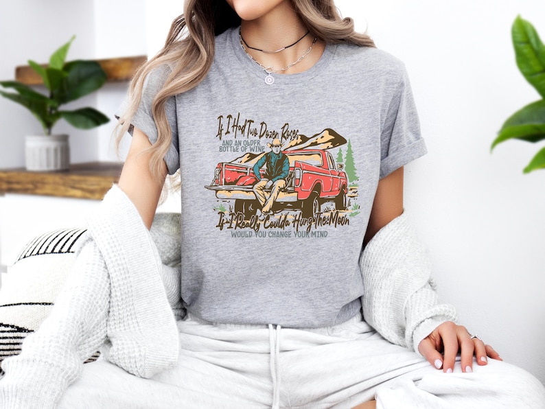 May include: Gray t-shirt with a vintage illustration of a man sitting in the back of a red pickup truck. The text on the shirt reads "If I Had Two Dozen Roses and an Older Bottle of Wine If I Really Could Hang the Moon Would You Change Your Mind".