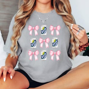 Op de afbeelding: Grijs T-shirt met een herhalend patroon van roze strikken en blikjes Sprite.