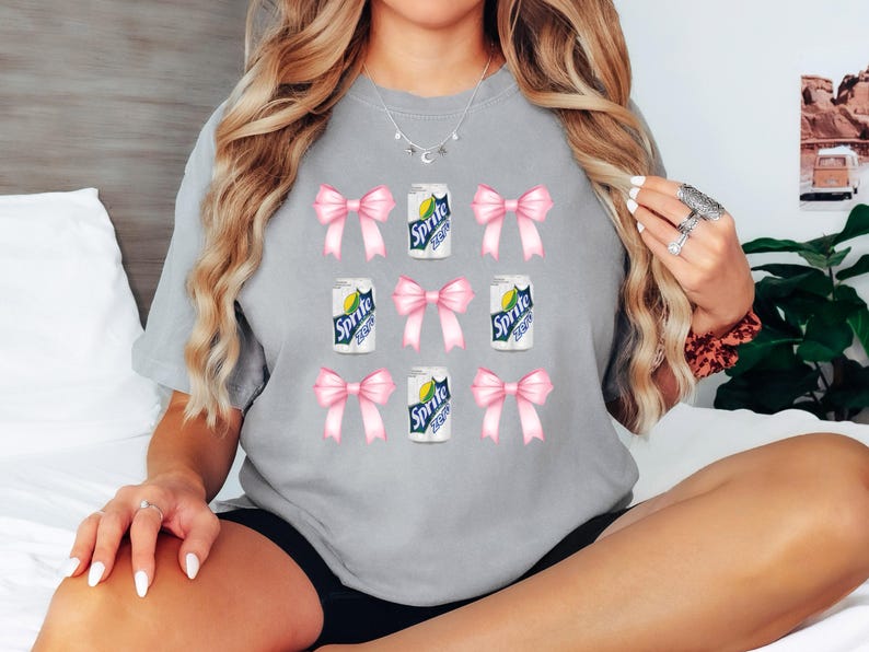 Op de afbeelding: Grijs T-shirt met een herhalend patroon van roze strikken en blikjes Sprite.