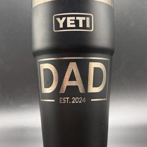 May include: Black Yeti tumbler with a gold inscription that reads "DAD EST. 2024".