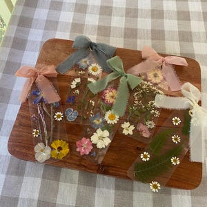 May include: Five clear bookmarks with pressed flowers and colorful ribbons. Each bookmark features a different assortment of dried flowers, including daisies, pansies, and ferns. The ribbons are in shades of pink, blue, green, and white. The bookmarks are displayed on a wooden board.