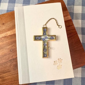 May include: A gold-toned cross-shaped bookmark with a floral design, attached to a chain, rests on a white book. The cross features blue and purple flowers. The book has a white cover and a gray spine. Gold flower illustrations are on the book.