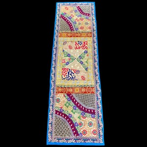 May include: A long, rectangular, decorative textile with a vibrant, patchwork design. The textile features a blue border and a central panel with geometric and floral patterns in red, yellow, green, and purple. The overall aesthetic is colorful and ornate.