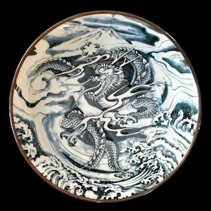May include: A round ceramic plate with a detailed illustration of a dragon in shades of blue and white. The dragon is surrounded by clouds and waves, with a mountain in the background. The plate has a brown rim.