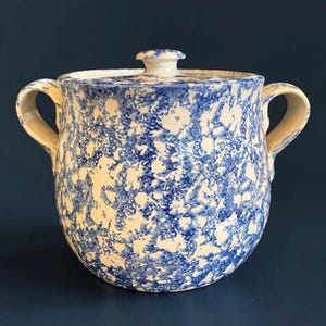 May include: A ceramic sugar bowl with a lid and two side handles, featuring a speckled blue and white sponge-painted pattern. The vessel has a rounded body and a matching lid with a small knob handle, showcasing a consistent mottled design.