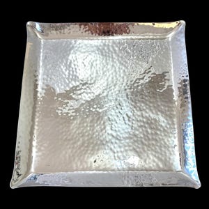 May include: A square, hammered silver tray with slightly curved edges. The tray has a textured surface and reflects light, creating a shimmering effect. The corners are gently rounded, and the overall design is simple and elegant. Ideal for serving or display.