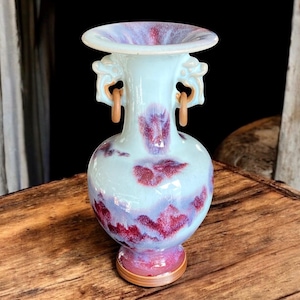 May include: A small, light blue and purple ceramic vase with two handles in the shape of stylized animal heads. The vase has a flared rim and a rounded body with a swirling pattern.