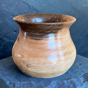 May include: A small, hand-turned wooden bowl with a smooth, polished finish. The vessel features a wide, flared rim and a rounded body, showcasing natural wood grain patterns that transition from a light tan base to a dark brown upper section.