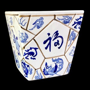 May include: A white ceramic planter with a geometric design. The planter is decorated with blue illustrations of birds, clouds, and Chinese characters. The edges are outlined in a gold color.