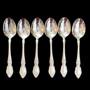 May include: Six silver spoons with ornate handles are arranged against a black background. The spoons have a reflective surface, and the handles feature a detailed floral design. The spoons are evenly spaced.