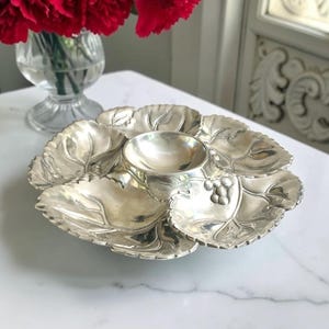 May include: A silver-toned metal serving dish shaped like a cluster of leaves arranged in a circle. The dish features a central circular bowl surrounded by six individual leaf-shaped compartments, with small embossed grape clusters on one leaf.