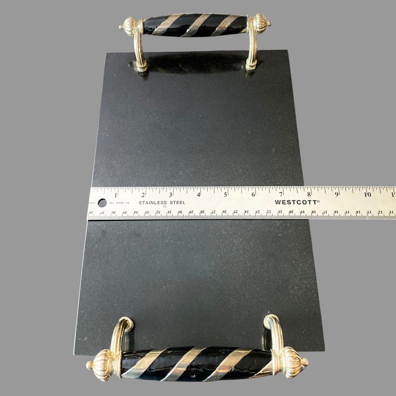 May include: A black serving tray with gold-coloured handles and black and gold striped accents. The tray is approximately 25 cm long. The handles have a decorative, ornate design.