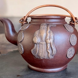May include: A brown clay teapot with a copper handle and decorative silver accents. The teapot features a raised design of a person in traditional clothing.