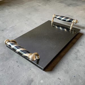 May include: A rectangular black serving tray with gold-coloured handles. The handles have a black and silver striped design. The tray is made of a solid, dark material, and the handles are ornate.